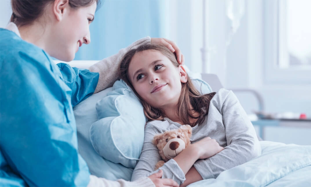 A new pediatric patient lies in a hospital bed, holding a teddy bear, while a nurse gently comforts her